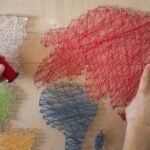 Hands creating a colorful string art map on a wooden board using vibrant threads.